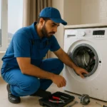 فني صيانة يرتدي زيّاً أزرق وقبعة يفحص غسالة تحميل أمامية في منزل، بجانبه صندوق أدوات وأجزاء.صيانة غسالات اوتوماتيك في مكة المكرمة